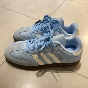 Adidas Samba ljusblå sneakers - Säljer ett par Adidas Samba sneakers i ljusblått med vita ränder och detaljer. Skorna har klassisk brun sula och snörning framtill. Perfekt för dig som gillar retrostil och vill sticka ut med en fräsch färg på fötterna. Prislapp finns kvar, aldrig använda.