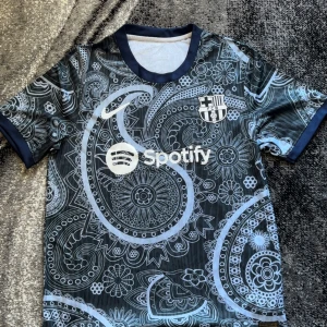 Barcelona fotbollströja  - Snygg Barcelona fotbollströja med Spotify-logga och paisleymönster i blått och svart. Kortärmad modell med klubbmärke på bröstet och rund halsringning. Perfekt för dig som vill sticka ut på planen eller på läktaren.