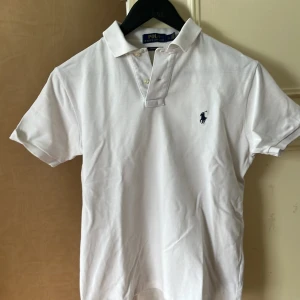 Ralph Lauren Piké vit - Klassisk vit pikétröja från Polo Ralph Lauren med blå broderad logga på bröstet. Tröjan har korta ärmar, krage och knappar framtill. Modellen är custom slim fit och passar perfekt till en stilren look.