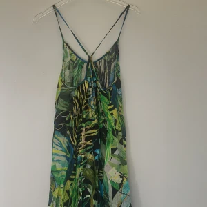 Grön långklänning med tropiskt mönster från H&M - Säljer en långklänning med tropiskt mönster i grönt, blått och gult från H&M. Klänningen har smala axelband, djup rygg och är tillverkad i ett tunt, luftigt material med 87% linocell. Perfekt för sommardagar!
