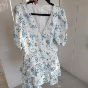Blommig kortklänning med volang - Superfin vit kortklänning med blått blommönster och volangdetaljer. Klänningen har v-ringning, puffiga korta ärmar och markerad midja. Perfekt för sommarens alla tillfällen! Köpt på vinted och hittar inget märke. Känns luftig och är ganska kort. Endast testad av mig, men den är lite liten. Byter gärna mot en liknande klänning i storlek S. Kan skicka fler bilder om så önskas. 😊