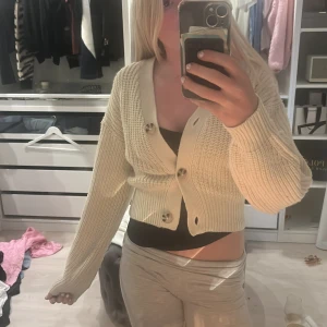 Beige croppad stickad kofta - Säljer en beige croppad stickad kofta med stora knappar framtill. Koftan har lång ärm och bred ribbad kant vid ärmslut och nederkant. Perfekt för lager-på-lager och enkel att styla till olika outfits.