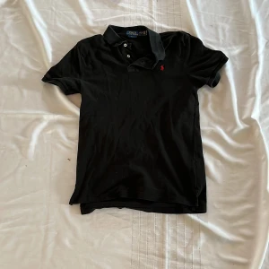 Svart pikétröja från Polo Ralph Lauren - Klassisk svart pikétröja från Polo Ralph Lauren med liten röd broderad logga på bröstet. Tröjan har korta ärmar, krage och knappar framtill. Perfekt för en stilren och tidlös look. Såklart äkta inga defekter.