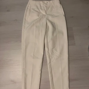 Vita kostymbyxor från Amisu - Snygga vita kostymbyxor från Amisu i storlek 34. Byxorna har hög midja, raka ben och pressveck framtill. Perfekta för en clean och stilren look. Stängs med knapp och dragkedja framtill.
