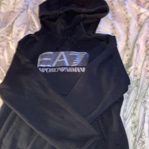Svart hoodie från Emporio Armani EA7 - Säljer en svart hoodie från Emporio Armani EA7 med stor logga i silver på bröstet. Tröjan har huva med dragsko och är tillverkad i mjukt material. Perfekt för en avslappnad och stilren look.