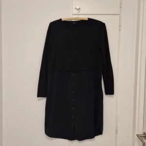 Mörkblå tvådelad klänning/tunika med knappar från COS - Mörkblå tvådelad klänning/tunika från COS med lång ärm och knappar framtill. Klänningen har en rak passform och är tillverkad i ett mjukt material. Enkel och stilren design som passar perfekt till många tillfällen. Liten lagning på höger sida, nacke.