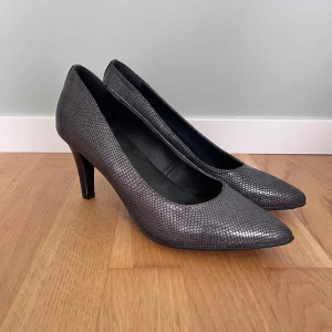 Grå pumps med ormskinnsmönster - Säljer ett par snygga grå pumps med spetsig tå och klack. Skorna har ett stilrent ormskinnsmönster och är perfekta för dig som vill ha något elegant och trendigt. Passar till många olika outfits och ger en lyxig känsla.
