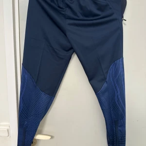 Mörkblå Brasilien Nike träningsbyxor - Säljer ett par mörkblå träningsbyxor från Nike med Brasilien-logga och turkosa detaljer. Byxorna har elastisk midja med dragsko och smal passform. Snyggt mönstrade partier på benen och dragkedjor vid bensluten.