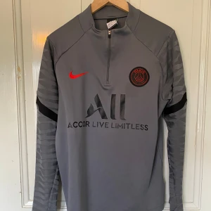 Nike x PSG träningströja - Säljer en grå långärmad PSG träningströja från Nike med svart och rött klubbmärke, röd Nike-logga och svart tryck. Tröjan har halv dragkedja och detaljerade ärmar med svarta inslag. Materialet är Dri-Fit för bra andningsförmåga.