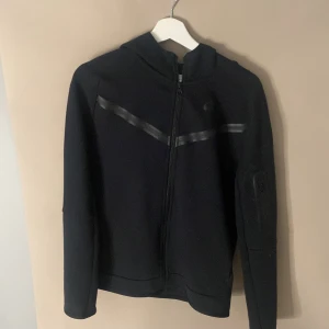 Svart hoodiejacka från Nike - Svart hoodiejacka från Nike med dragkedja framtill och diskreta svarta detaljer över bröstet. Jackan har huva och långa ärmar, perfekt för en sportig och avslappnad stil. 