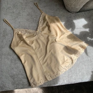 Beige linnetopp med spetsdetaljer - Säljer en beige linnetopp med tunna axelband och vacker spets längs urringning och nederkant. Toppen har en mjuk och glansig yta som ger en lyxig känsla. Perfekt att styla till olika outfits!