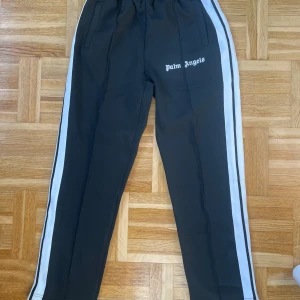  Palm Angels byxor i svart - Svarta träningsbyxor från Palm Angels med vita sidoremsor och logga tryckt på vänster lår. Byxorna har elastisk midja och raka ben. Perfekta för en sportig look.