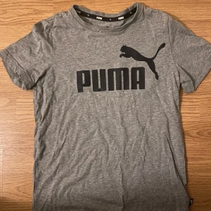 Grå t-shirt från Puma - Säljer en grå t-shirt från Puma med klassisk logga tryckt i svart på bröstet. T-shirten har rund halsringning och korta ärmar. Perfekt för en sportig och avslappnad stil. Kostar bara 50kr för en extrem bra t-shirt! Är storlek 152 (11-12 år) både för tjejer och killar! Pris kan diskuteras.
