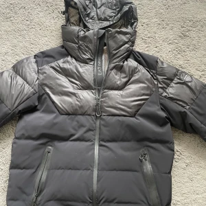 Svart pufferjacka från EA7 Emporio Armani - Säljer en svart pufferjacka från EA7 Emporio Armani med huva och dragkedja. Jackan har två fickor med dragkedja och EA7-logga på ärmen. Den har även två innerfickor i nät. Luvan går att veckla in. Perfekt för kyliga dagar och riktigt snygg streetstil. (Äkta)