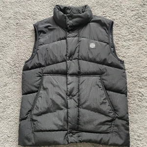 Svart dunväst från Stone Island - Säljer en svart dunväst från Stone Island med hög krage och knappar framtill. En dragkedja finns även under knapparna. Västen har två stora fickor på framsidan och klassisk Stone Island-logga på bröstet. Perfekt för lager-på-lager under kyliga dagar. Använd ett fåtal gånger och är i ett super skick. (Äkta)