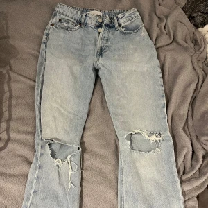 Ljusblå slitna jeans från HM - Säljer ett par ljusblå jeans med raka ben och stora hål på båda knäna. Jeansen har knappar i gylfen och klassisk femficksdesign. Perfekta för en avslappnad och trendig look.