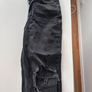 Svarta slitna jeansbyxor - Säljer ett par svarta jeansbyxor med slitningar och hål vid knäna. Byxorna har klassisk femficksdesign, dragkedja och knapp framtill. Perfekta för en avslappnad och trendig stil.