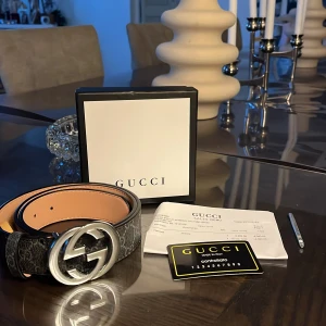 Gucci bälte  - Säljer ett svart bälte från Gucci med klassiskt GG-mönster och stort, rundat spänne i silver. Kommer med originalask och certifikat. Äkta och byten funkar också.