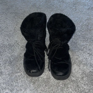 Svarta fluffiga vinterboots - Säljer ett par svarta boots med fluffigt pälsfoder och snörning framtill. Skorna har en högre skaft och en rejäl platt sula som passar perfekt för kalla dagar. De är både snygga och varma, perfekta för vintern.