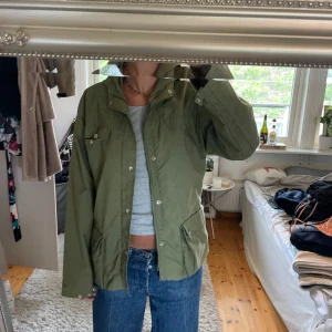 Grön vindjacka från Fjällräven - Snygg grön vindjacka från Fjällräven vintage med huva och flera fickor framtill. Jackan har knappar och dragkedja samt klassisk Fjällräven-logga på ärmen. Perfekt för utomhusaktiviteter och enkel att matcha. Storlek M