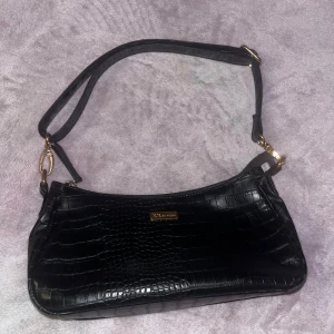 Svart axelväska  - Snygg svart axelväska. Väskan har guldfärgade detaljer och en justerbar axelrem. Ganska rymlig med innerficka med dragkedja. Lädret undertill är lite nött då den varit använd till vardags tidigare. Säljs för 40kr+frakt 