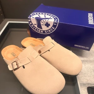 Beige Birkenstock Boston tofflor i mocka - Säljer ett par klassiska Birkenstock Boston i beige mocka. Skorna har en justerbar rem med spänne och en bekväm fotbädd. Oanvända! perfekt för stranden!