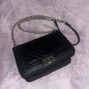 Svart axelväska med kedja - Säljer en svart axelväska med quiltat mönster och kedjerem. Väskan har ett snyggt spänne framtill, är rymlig med en liten ficka innnuti och designnen passar till många tillfällen. Säljs för 180kr+frakt 