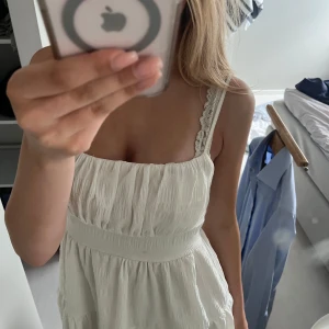 Vit somrig klänning med volang - Säljer en söt vit klänning med smala axelband och volangdetaljer. Klänningen har en luftig passform och är tillverkad i ett lätt material, perfekt för varma dagar. Axelbanden har spetsdetaljer som ger en extra fin touch.