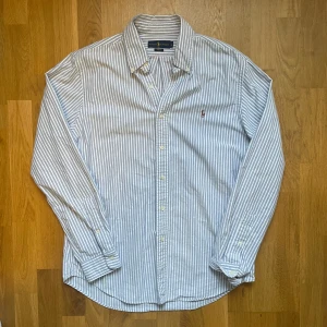 Ralph lauren skjorta  - Säljer denna Snygga vit och ljusblå randiga skjorta från Ralph lauren, den är i mycket bra skick och har inga fläckar eller missfärgningar, Storlek L och modell slim fit den är 78 cm lång och 54 cm bred, hör gärna av dig vid frågor!