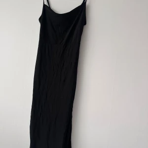Svart långklänning med smala axelband - En stilren svart långklänning med smala axelband och rak passform. Klänningen har en enkel design och är tillverkad i ett mjukt, lätt material som faller snyggt längs kroppen. Perfekt för dig som gillar minimalistisk stil.