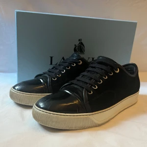 Svarta sneakers från Lanvin - Snygga svarta sneakers från Lanvin med vit platt sula och rund tå. Skorna har snörning och är tillverkade i blankt skinn som ger en lyxig still. Skorna är i 9/10 skick✅ hör av er vid frågor 😀 box och dustbag fås med 