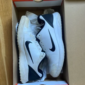 Nike Infinity G Golfskor vita och svarta - Nike Infinity G golfskor i vitt och svart med klassisk Nike-logga på sidan. Skorna har snörning, rund tå och platt sula. Perfekta för golfbanan med sportig och stilren design. De är lite smutsiga men det går att få rena om man vill, endast använda under en säsong.