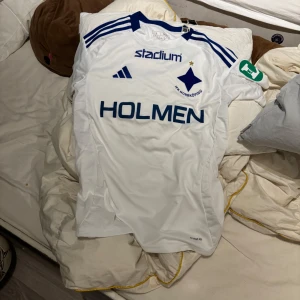 IFK Norrköping vit matchtröja - Säljer en vit IFK Norrköping fotbollströja från Adidas med blå detaljer och klubbmärke på bröstet. Tröjan har korta ärmar, blå ränder på axlarna och sponsorloggor på framsidan. Perfekt för dig som vill visa ditt stöd på läktaren. Jag köpte den men den var för liten. Den är ny och aldrig använd