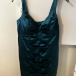 Mörkgrön satin klänning från Ginatricot  - Snygg mörkgrön klänning i satin från NLY One. Klänningen har breda axelband, figurnära passform och dekorativa veck framtill. Perfekt för dig som vill sticka ut med en elegant look.