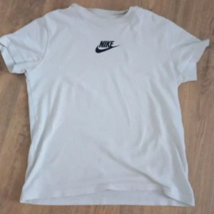 Vit t-shirt från Nike  - Säljer en klassisk vit t-shirt från Nike med svart logga på bröstet. T-shirten har rund hals och korta ärmar. Perfekt för en sportig och avslappnad stil.
