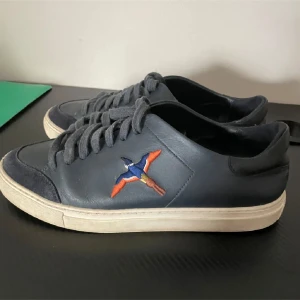 Mörkblå sneakers Axel Arigato   - Snygga mörkblå sneakers från Axel Arigato med en färgglad broderad fågel på sidan. Skorna har snörning och vit platt sula. Perfekta för dig som vill ha en stilren men unik look. Kan sänka pris vid snabb affär 
