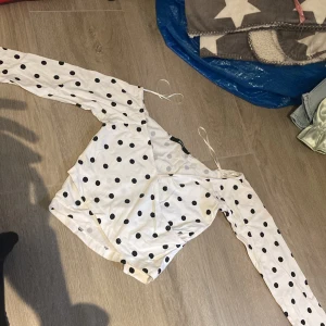 Vit prickig omlottopp från Bik Bok - Säljer en vit omlottopp från Bik Bok med svarta prickar. Toppen har långa ärmar och knytband vid axlarna. Perfekt för dig som gillar trendiga och lekfulla mönster!