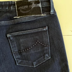 Mörkblå jeans från Jacob Cohën - Snygga mörkblå jeans från Jacob Cohën med klassisk femficksdesign och dekorativa sömmar på bakfickorna. Jeansen är i en rak modell och har silverfärgade knappar framtill. Tillverkade av japanskt tyg med italienskt hantverk.