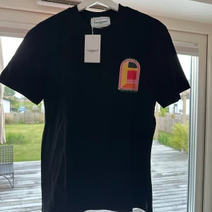 T shirt Casablanca  - Svart t-shirt från Casablanca med färgglatt grafiskt tryck på bröstet och stor illustration med texten 'Casablanca' på ryggen. Klassisk passform och rund halsringning. Perfekt för dig som gillar stilrena plagg med en twist. Oanvänd, storlek M passar S 