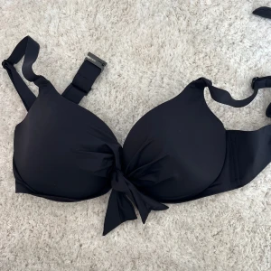Svart bikiniöverdel med rosett - Snygg svart bikiniöverdel med vadderade kupor och en rosett framtill. Justerbara axelband och spänne i ryggen för perfekt passform