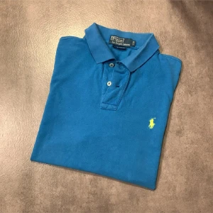 Blå pikétröja från Polo Ralph Lauren - Säljer en klassisk blå pikétröja från Polo Ralph Lauren med gul broderad logga på bröstet. Tröjan har korta ärmar, krage och knäppning framtill. Perfekt för en stilren och avslappnad look. Tröjan ör smutsig på några ställen som ni ser på bild 5. Bara att höra av er vid frågor eller funderingar! 😀