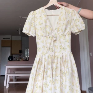 Blommig kortklänning med puffärm och knyt - Söt kortklänning med ljusgul blommig print på vit botten. Klänningen har puffärmar, v-ringning och knyt framtill. Kjolen är utställd och materialet känns lätt och somrigt. Perfekt för varma dagar och midsommarfirande.