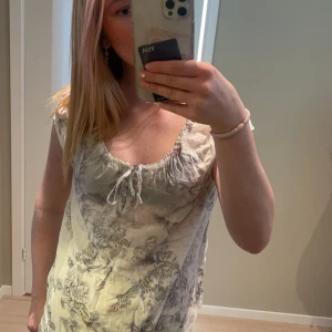 Vit blus med mönster och knytning - Säljer en söt vit blus med vitt/grått och fjärilsmönster. Blusen har korta ärmar, rundad halsringning med knytning framtill och är tillverkad i ett tunt, luftigt material. Finns inte längre och köpa, Perfekt för sommaren!