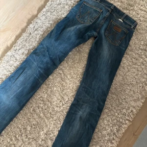 Blå jeansbyxor från Wrangler - Säljer ett par klassiska blå jeansbyxor från Wrangler med bootcut ben och snygga kontrastsömmar. Byxorna har fem fickor och tydliga W-detaljer på bakfickorna. Perfekta för dig som gillar tidlös denimstil.
