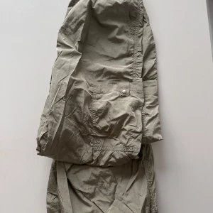 Beiga cargopants från H&M Divided - Säljer ett par beiga cargopants från H&M Divided i storlek XS. Byxorna har flera fickor, dragsko i midjan och är tillverkade i ett lätt syntetmaterial. Perfekta för en avslappnad och trendig stil.