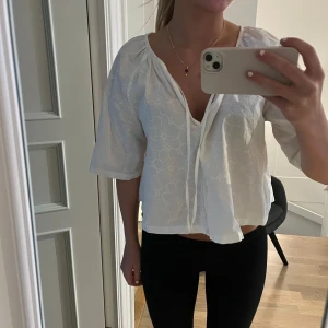 Vit topp - Säljer en söt vit blus med diskret blommigt mönster och knyt vid halsen. Blusen har trekvartsärm och en luftig, lite lös passform. Perfekt till vår och sommar!