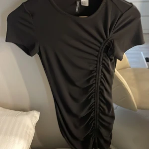 Svart kortklänning med rynk från H&M Divided - Svart kortklänning från H&M Divided med korta ärmar och rund halsringning. Klänningen har en snygg rynkdetalj längs ena sidan som ger en cool och modern look. Perfekt för dig som gillar enkel men trendig stil.