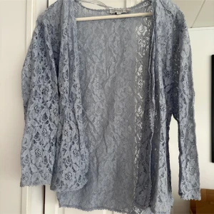 Ljusblå spetskofta från Flash Woman - Säljer en ljusblå, långärmad spetskofta från Flash Woman. Koftan är tunn och genombruten med blommigt spetsmönster och öppen framtill. Perfekt för att lägga till en extra touch till din outfit.
