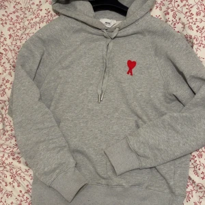 Grå Ami Hoodie - Säljer en stilren grå hoodie från Ami med röd broderad logga (hjärta och A) på bröstet. Perfekt för en avslappnad och trendig look.