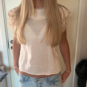 Vit blus med volangärmar och spetsdetaljer - Söt vit blus med korta volangärmar och fina spetsdetaljer längs ärmar och halsringning. Blusen har en lös passform och är tillverkad i ett tunt, luftigt material. Perfekt för varma dagar och ger en romantisk känsla.